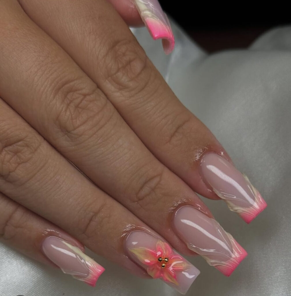 Une main avec de longs ongles carrés en acrylique avec des pointes françaises roses, des accents de marbre blanc et jaune, et une fleur rose en 3D avec une perle dorée sur l'annulaire. Les ongles ont une finition brillante.