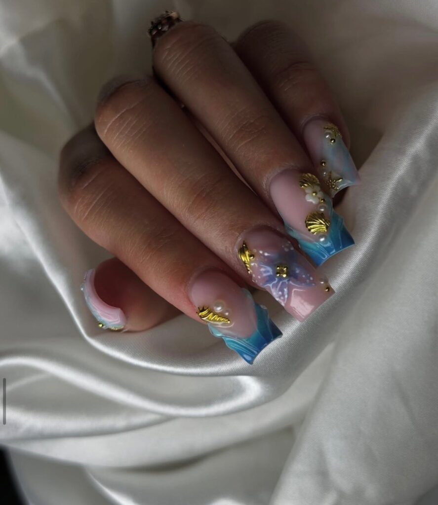 Une main aux ongles longs et carrés, ornée d'un motif ombré bleu et rose, de coquillages et d'étoiles de mer dorés, de fleurs blanches en 3D et d'ornements dorés, reposant sur un tissu de satin blanc.