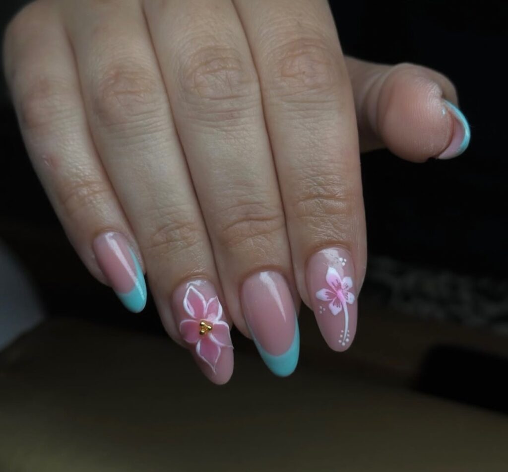 Une main aux ongles en forme d'amande peints dans une teinte rose pâle, avec des pointes bleues et de délicats motifs floraux sur deux ongles, dont l'un avec une fleur rose en 3D et un centre en perles d'or.