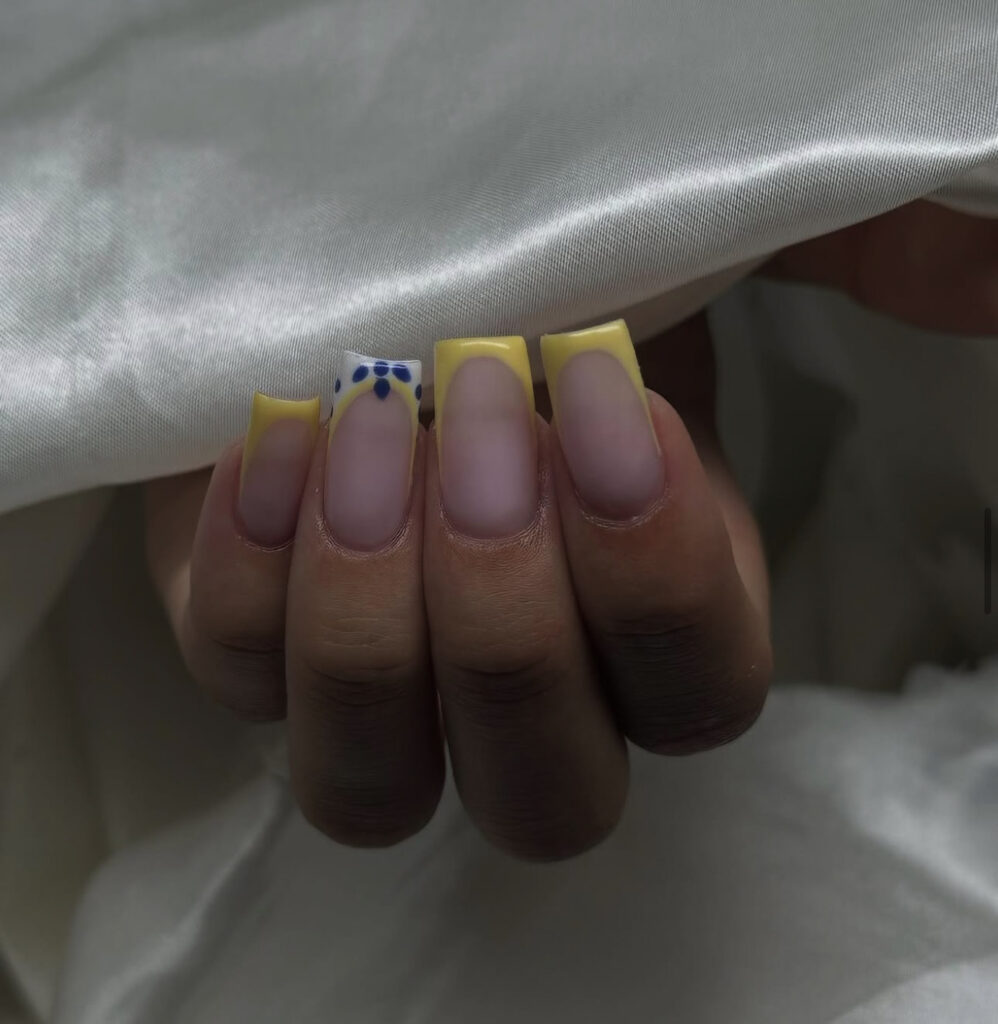 Une main avec des ongles de forme carrée présentant une manucure à la française de couleur jaune. L'un des ongles est orné de petits motifs floraux bleus et blancs près de la pointe. La main est partiellement enveloppée dans un tissu de satin blanc.