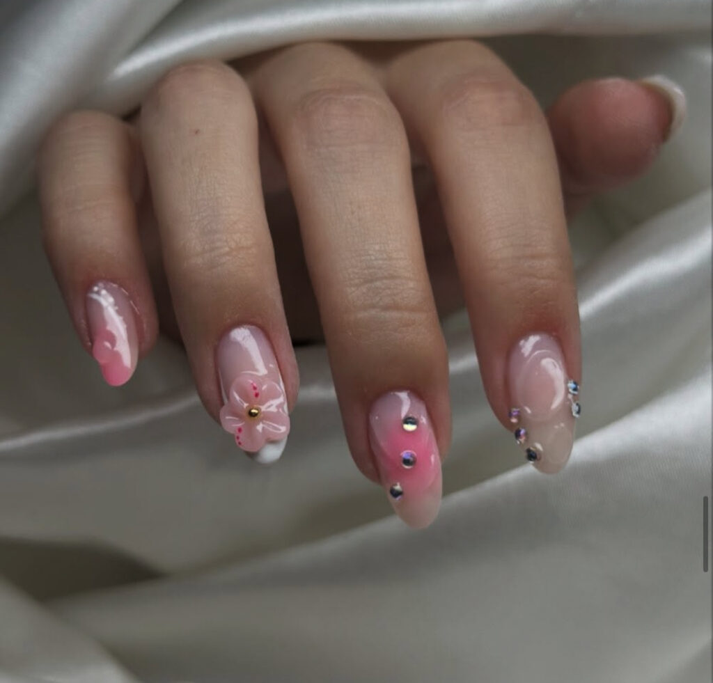 Une main aux ongles longs et en forme d'amande, avec un vernis ombré rose et nude, des accents de strass et un motif floral en 3D sur l'annulaire, le tout sur un fond de tissu blanc soyeux.