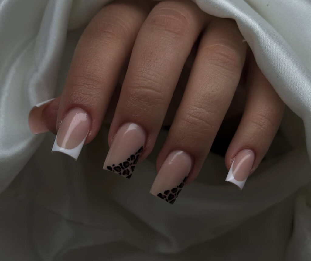Une main avec des ongles acryliques nus de forme carrée, avec des pointes françaises blanches sur la plupart des doigts et des accents de léopard noir près des pointes de deux ongles, reposant sur un tissu de satin blanc.