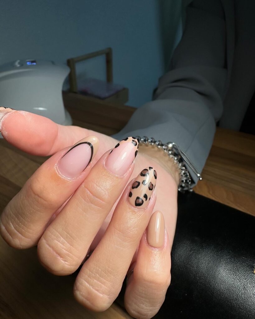 Une main avec des ongles manucurés présentant un mélange de motifs : deux ongles ont une base nude avec des taches de léopard noires et brunes, un ongle a une base nude avec une fine pointe noire, et deux ongles sont peints d'une couleur nude unie.