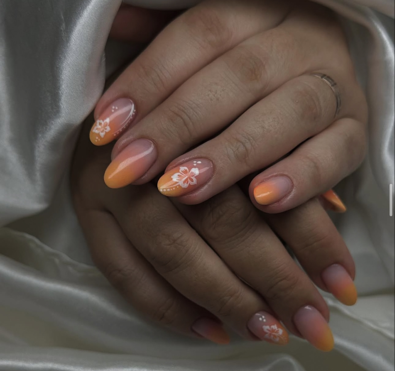 Mains aux ongles mi-longs en forme d'amande, présentant un motif ombré orange et jaune, des accents floraux blancs et de minuscules points blancs, reposant sur un tissu de satin blanc.