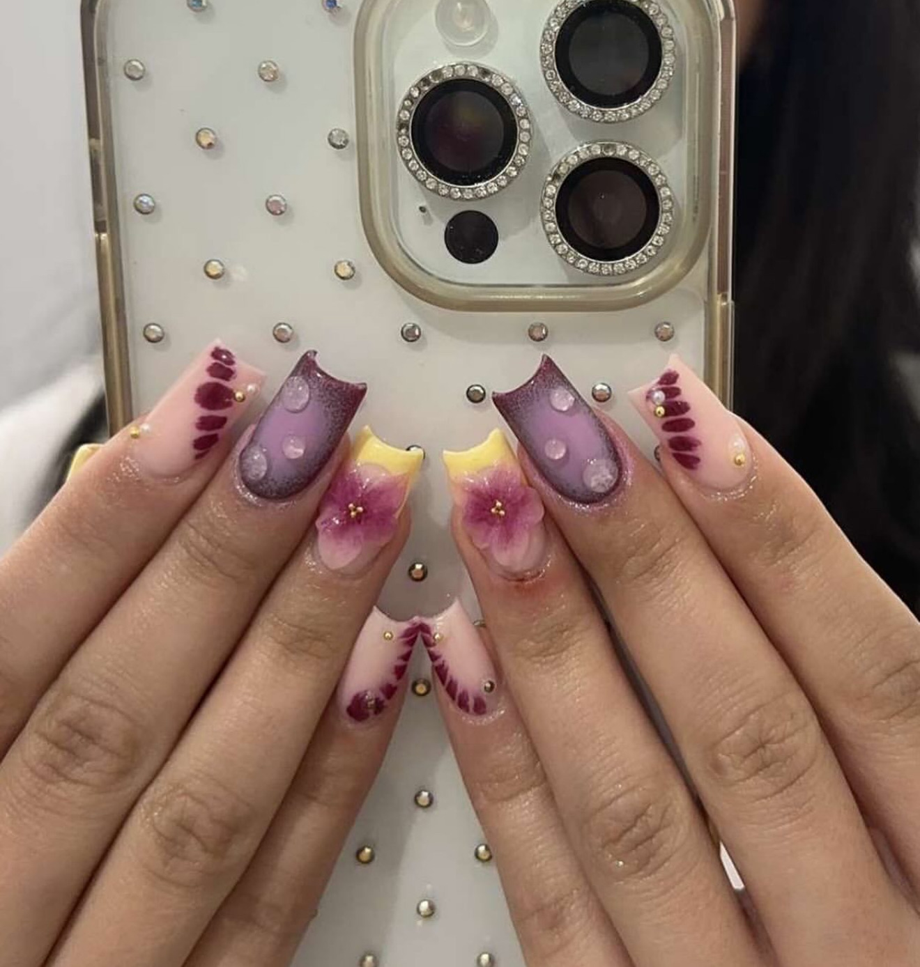 Gros plan sur des mains aux ongles longs et artistiques avec des pointes violettes et jaunes, des motifs floraux, des effets de gouttes d'eau en 3D et des accents de points, tenant un téléphone avec un étui orné de bijoux et de pois.