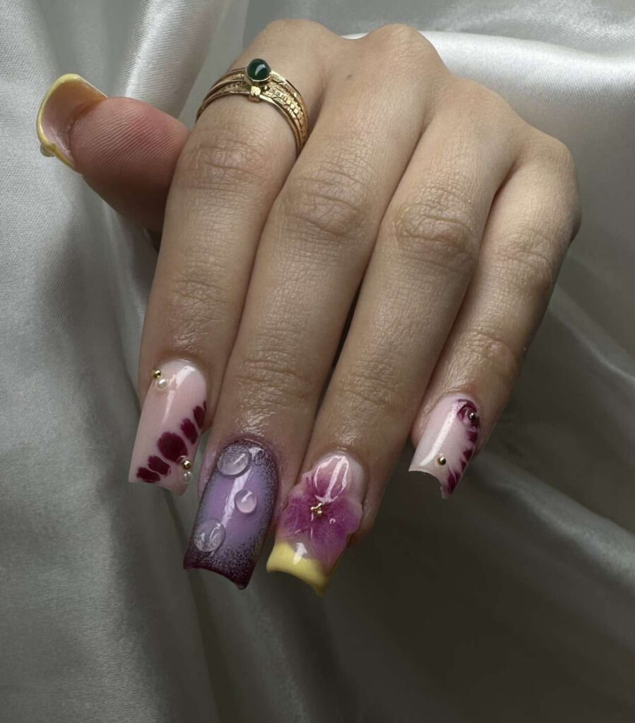 Une main aux ongles manucurés présentant un nail art complexe : tons roses et violets, effets de gouttes d'eau en 3D, fleur en 3D, clous dorés et motifs abstraits. La personne porte deux bagues en or sur un fond de tissu satiné.