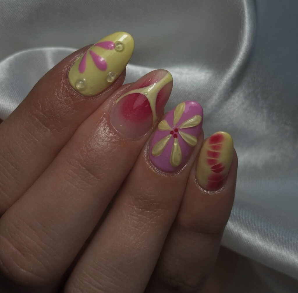 Une main aux ongles longs, en forme d'amande, ornés d'un nail art jaune et rose pastel, comprenant des motifs floraux, des pointes dégradées et de petites perles, reposant sur un tissu de satin blanc.