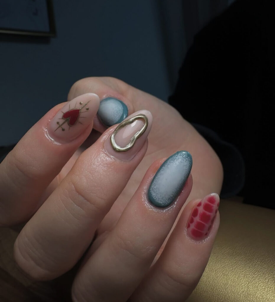 Une main au nail art créatif : un ongle est orné d'un cœur rouge avec des flèches dorées, un autre d'un accent ovale doré, deux ont des pointes ombrées bleues et un autre présente un motif rouge en peau de crocodile.