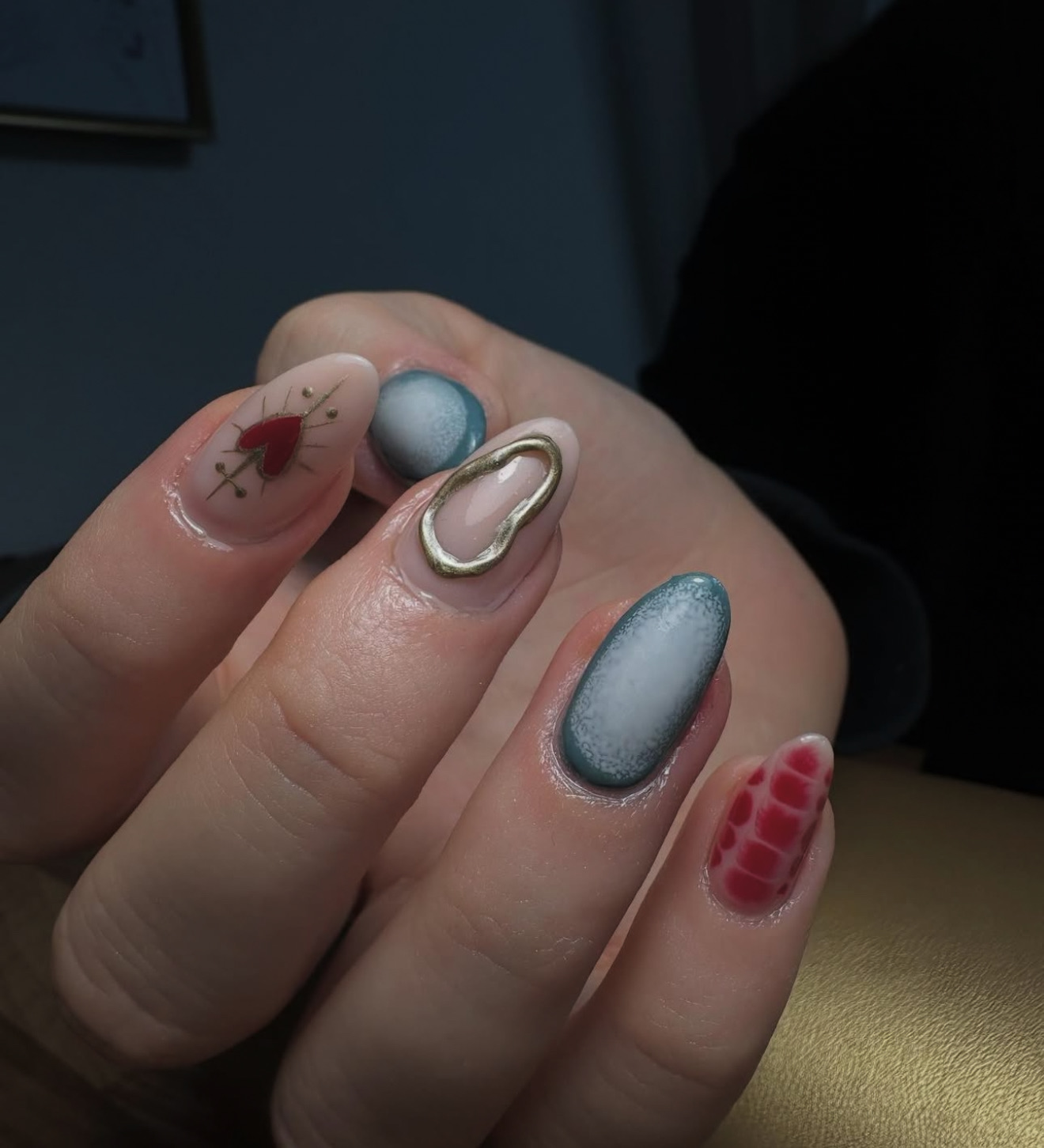 Une main au nail art créatif : un ongle est orné d'un cœur rouge avec des flèches dorées, un autre d'un accent ovale doré, deux ont des pointes ombrées bleues et un autre présente un motif rouge en peau de crocodile.