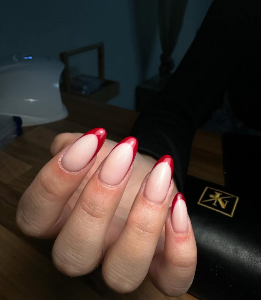Une main aux ongles en forme d'amande présente une base nue et des pointes rouges vibrantes, présentées sur un fond sombre avec un portefeuille noir et une surface en bois visible.