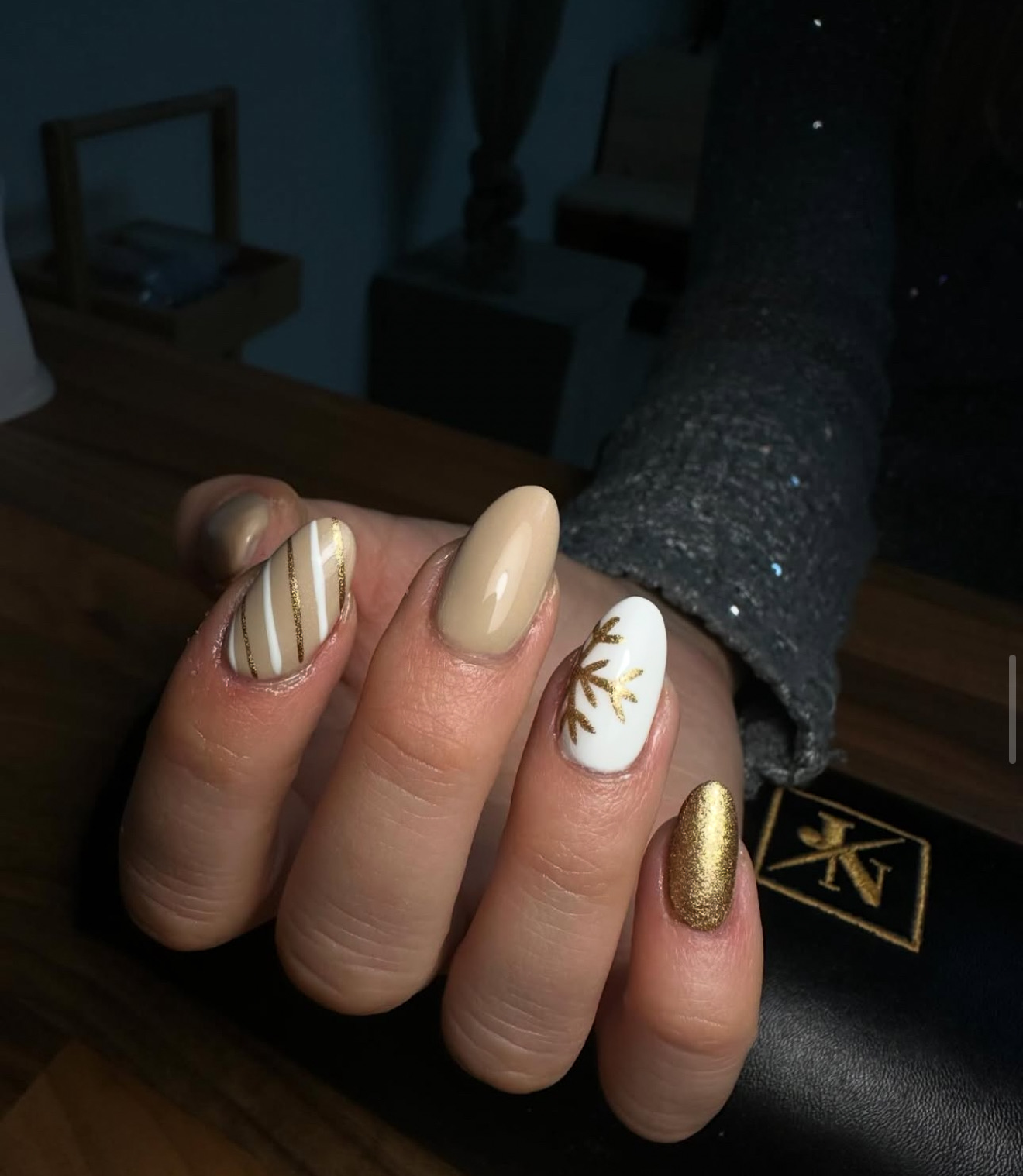 Une main avec des ongles en forme d'amande présentant un motif or et nude : des rayures dorées sur l'index, un nude solide sur le majeur, une feuille d'or sur fond blanc pour l'annulaire et de l'or métallique sur l'auriculaire.