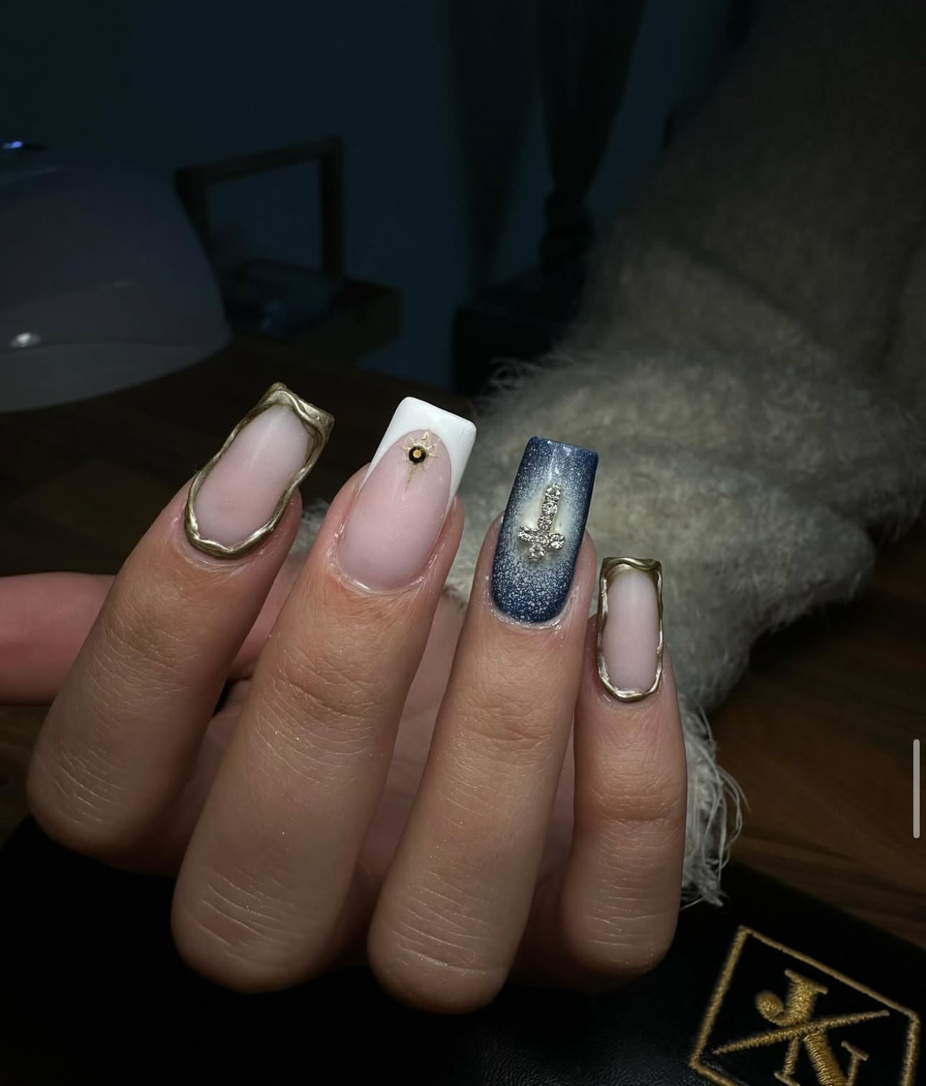 Une main avec de longs ongles de forme carrée présentant différents motifs : des contours métalliques dorés, un ongle blanc avec un clou doré et un ongle bleu avec un motif en forme de croix argentée étincelante.