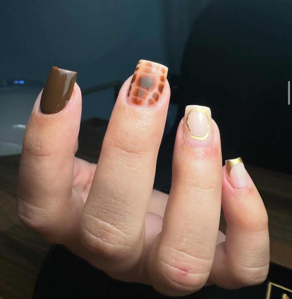 Une main avec des ongles manucurés : l'un peint en brun uni, l'autre avec un motif animalier brun, l'un avec un contour doré et l'autre avec une pointe dorée, tous dans des styles différents.