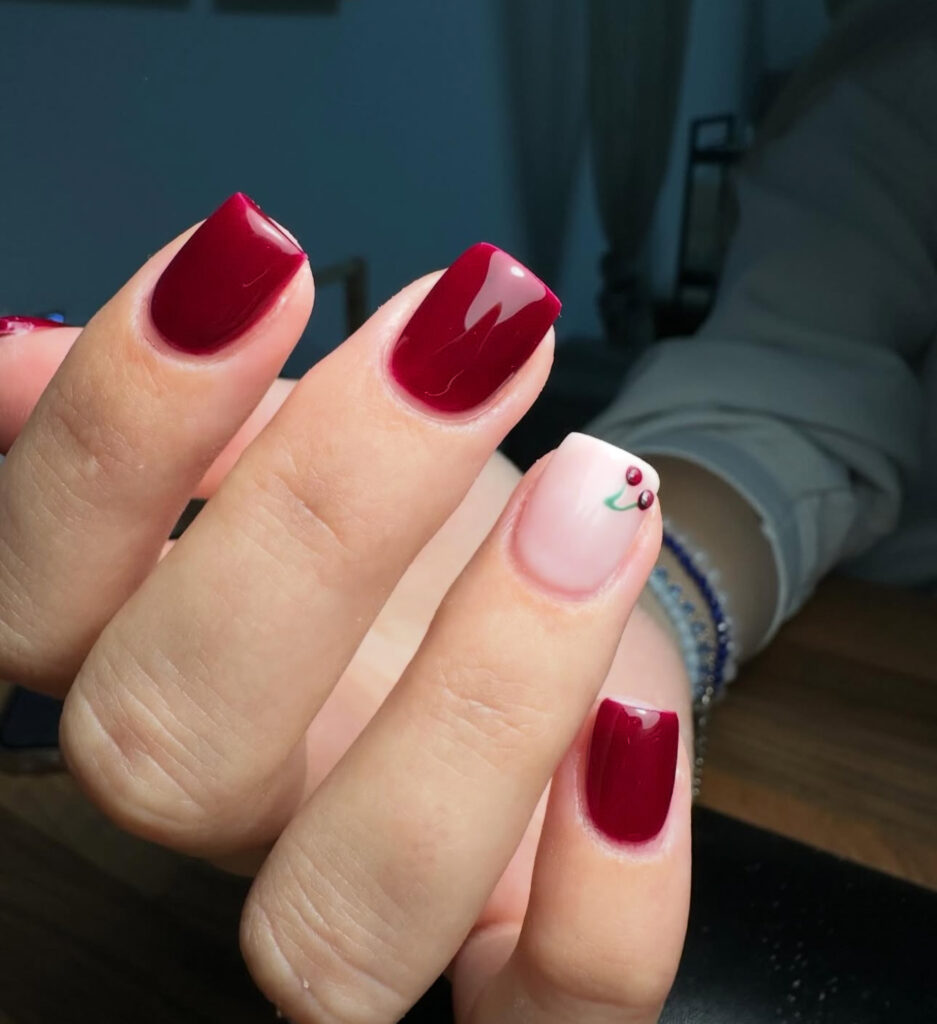 Une main aux ongles manucurés ; la plupart des ongles sont peints en rouge brillant, tandis que l'annulaire est peint en rose clair avec un petit motif de baie de houx et de feuille.