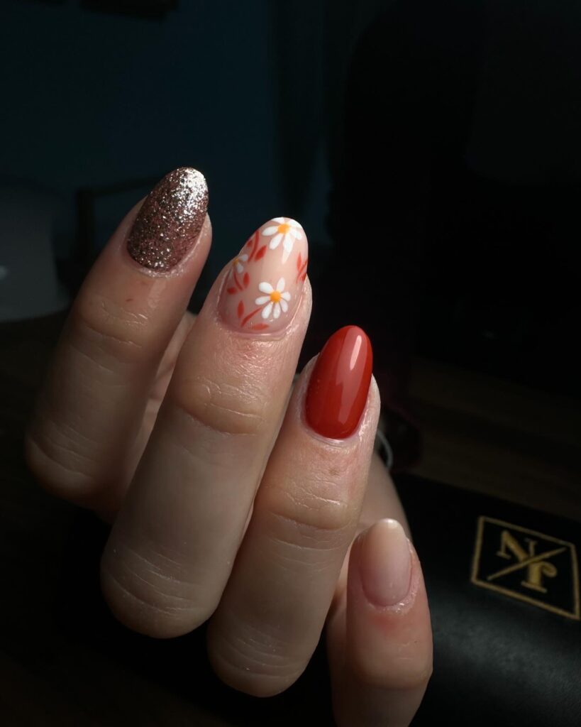 Une main dont les ongles sont peints dans différents styles : bronze pailleté, motif floral avec des fleurs orange et blanches, rouge-orange brillant et nude naturel. La main est tenue sur un fond sombre.