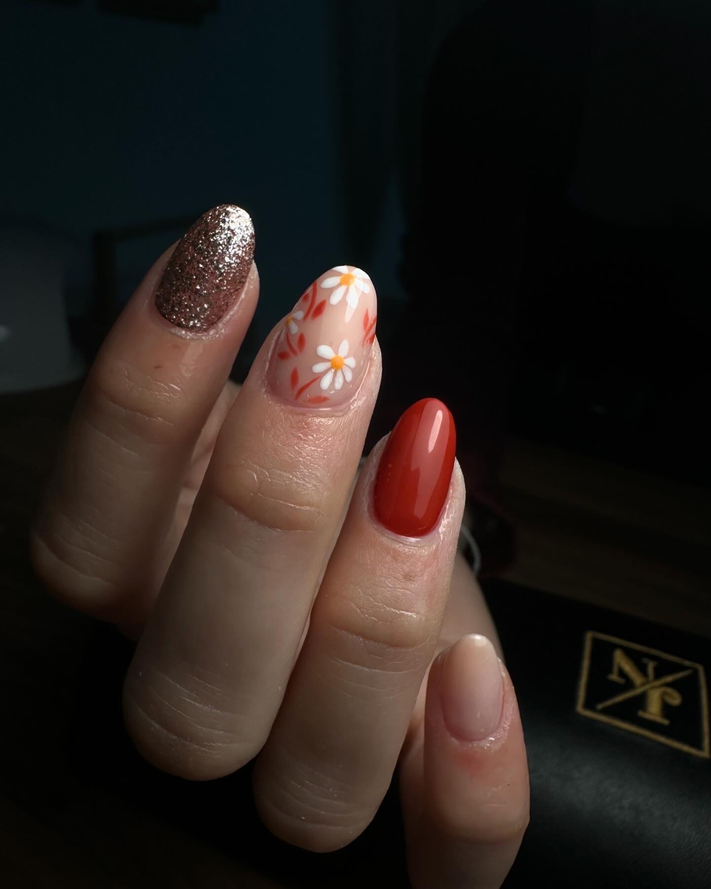 Une main dont les ongles sont peints dans différents styles : bronze pailleté, motif floral avec des fleurs orange et blanches, rouge-orange brillant et nude naturel. La main est tenue sur un fond sombre.