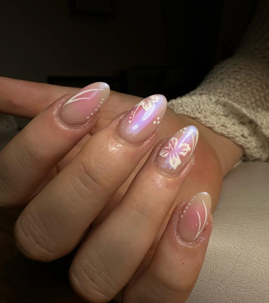 Une main avec des ongles en forme d'amande présentant un motif dégradé rose et blanc, un nail art floral et papillon blanc, et des accents délicats en forme de points. La personne porte une manche en tricot de couleur crème.