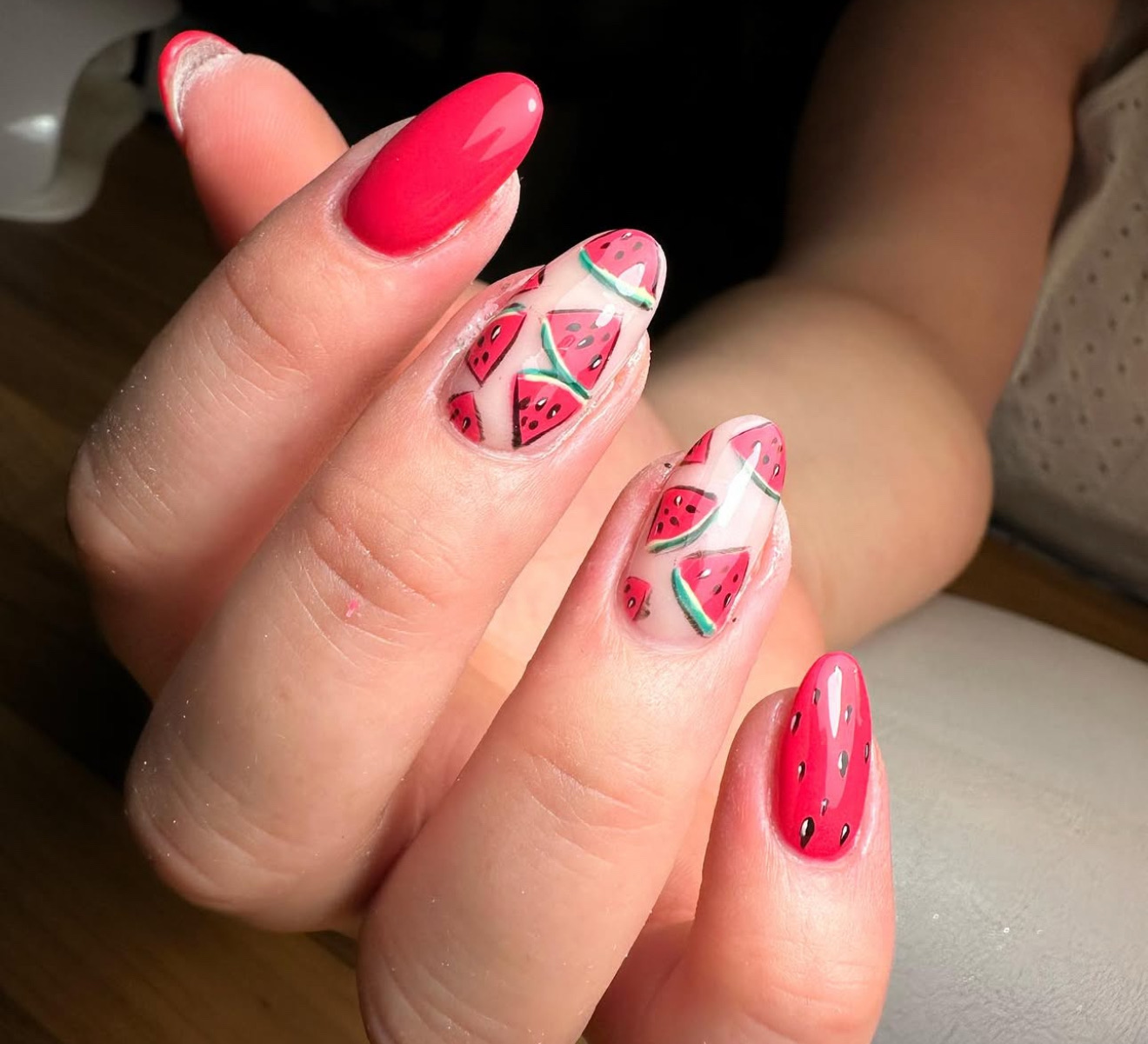 Une main avec des ongles manucurés sur le thème de la pastèque ; certains ongles ont un vernis rose avec des graines noires, tandis que d'autres présentent des tranches de pastèque avec des détails roses, verts et blancs.