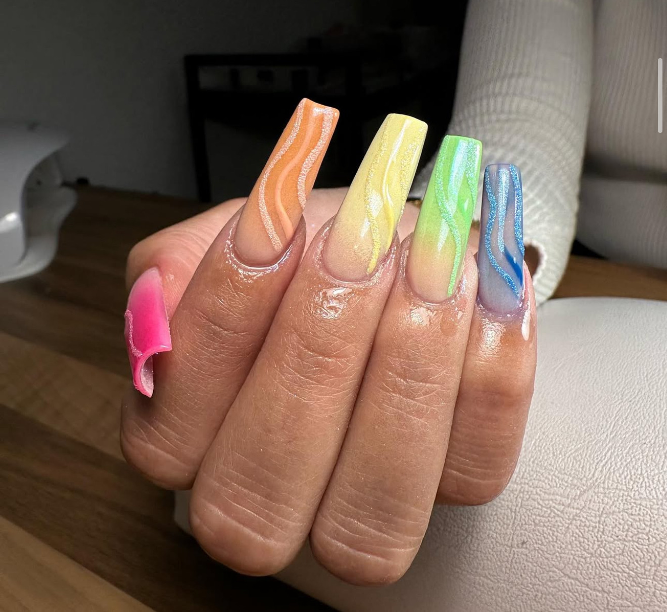Une main avec de longs ongles carrés en acrylique, chacun peint dans une couleur vive différente - rose, orange, jaune, vert et bleu - avec des motifs blancs ondulés sur chaque ongle. L'arrière-plan comprend une surface en bois et un coussin blanc.
