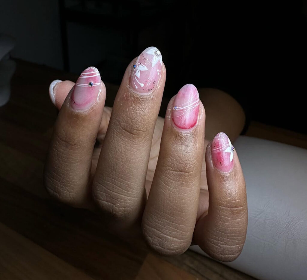 Une main aux ongles manucurés avec du vernis rose et blanc, des lignes argentées, de petits strass et un papillon blanc sur l'ongle du milieu, reposant sur une surface en bois.