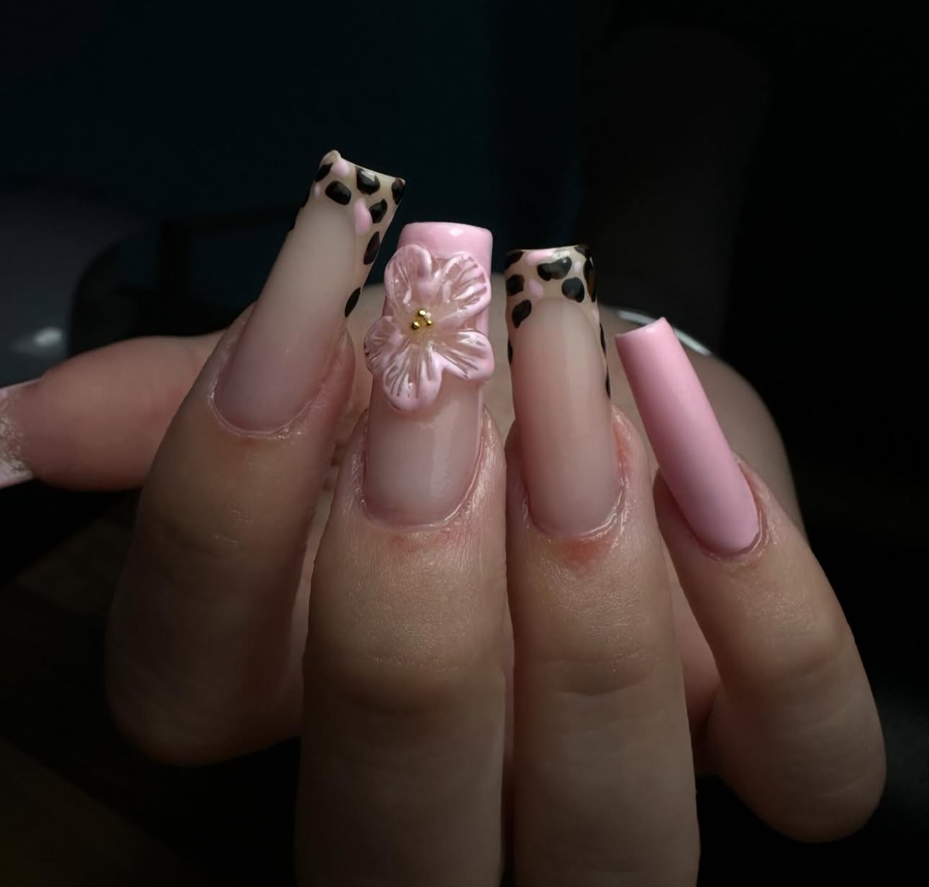 Une main avec de longs ongles carrés en acrylique présentant un motif ombré rose et nude, deux ongles avec des pointes en imprimé léopard, un ongle avec une fleur rose en 3D, et un ongle peint en rose uni.