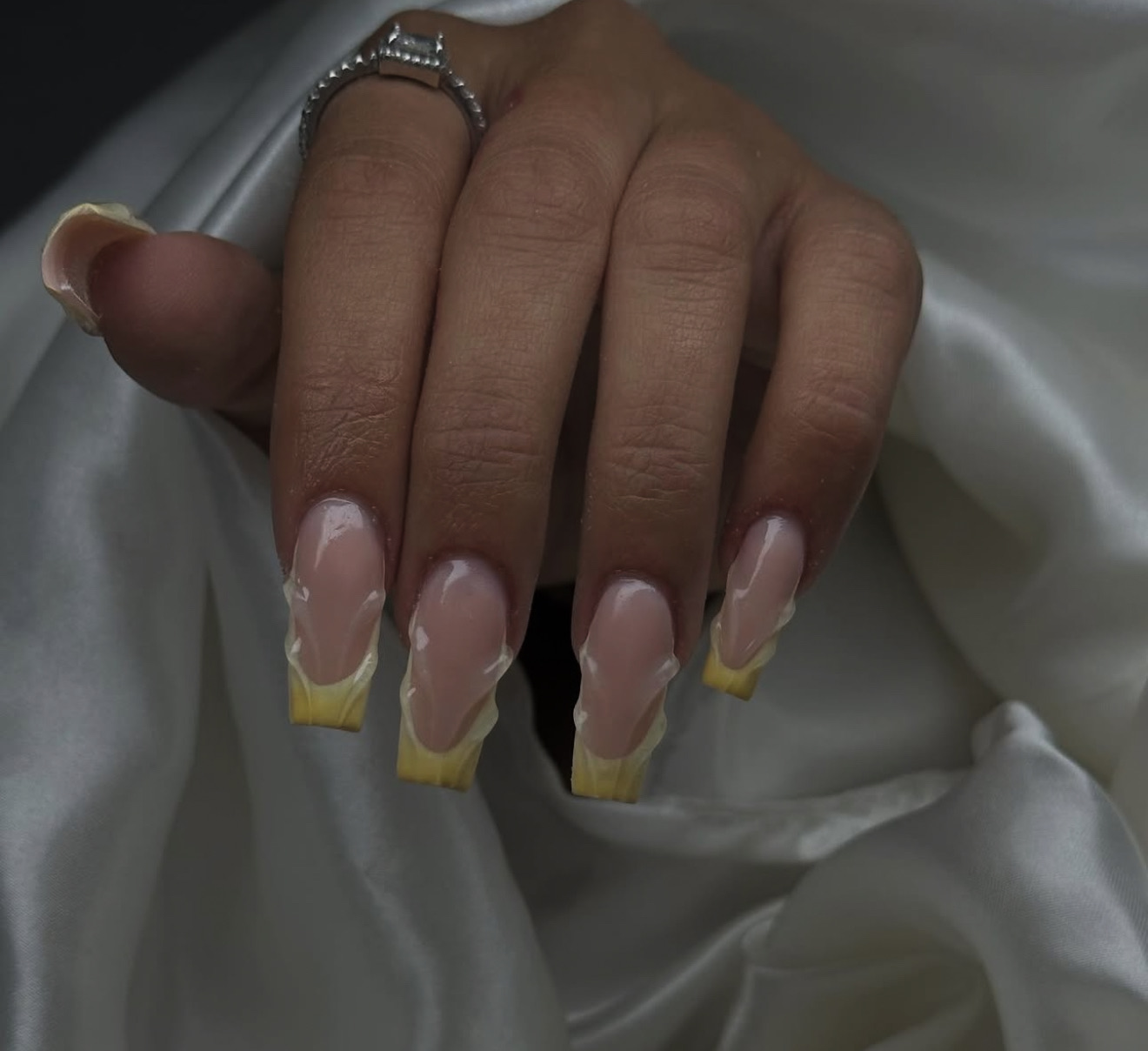 Une main avec de longs ongles acryliques de forme carrée ressemblant à des épis de maïs, reposant sur un tissu de satin blanc. Les ongles ont une pointe jaune et un dessin texturé imitant les grains de maïs.