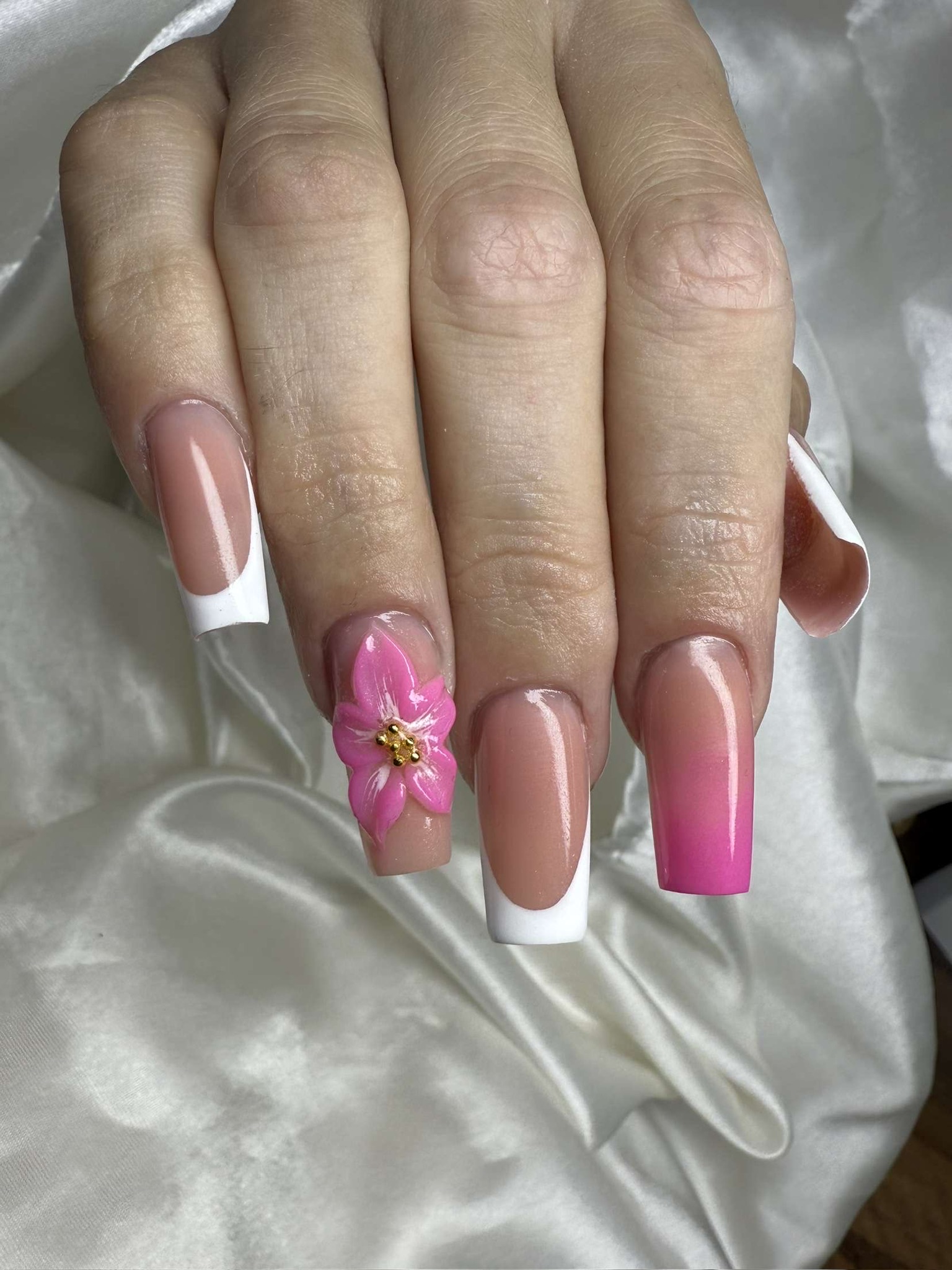 Une main aux ongles longs et carrés présente une manucure française, un ongle d'accentuation rose et un ongle décoré d'une fleur 3D rose détaillée et d'un centre doré, sur un fond de satin blanc.