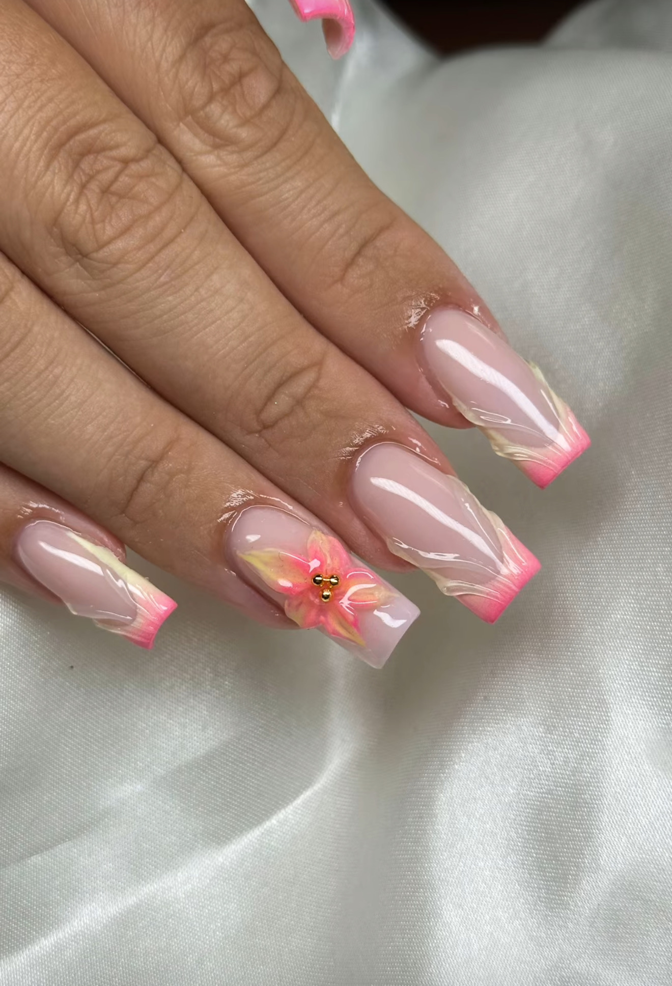 Une main avec de longs ongles acryliques de forme carrée présentant un motif French tip rose et blanc, des accents blancs et une fleur rose en 3D avec une petite pierre précieuse sur l'annulaire, le tout sur un fond blanc satiné.