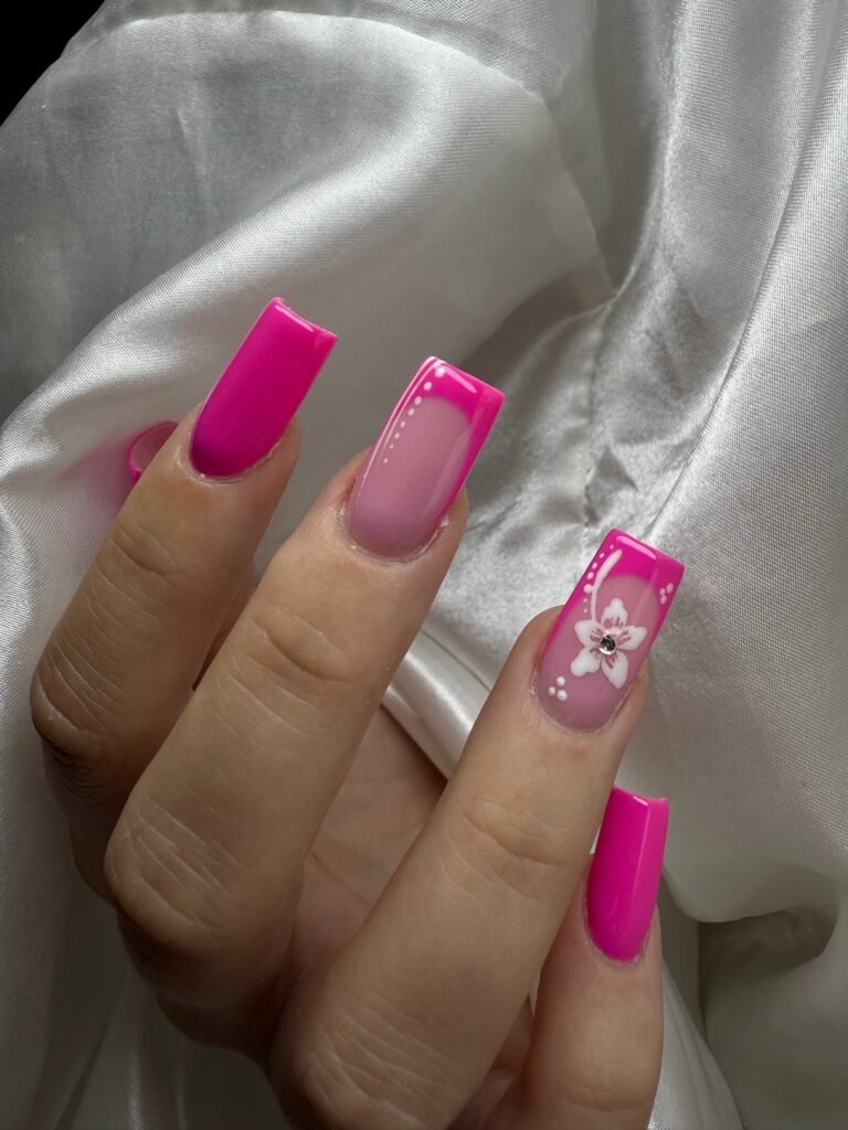 Une main aux ongles longs et carrés est recouverte d'un vernis rose vif. L'annulaire est orné d'un ongle clair avec une fleur blanche peinte et des points décoratifs. La main repose sur un tissu blanc soyeux.