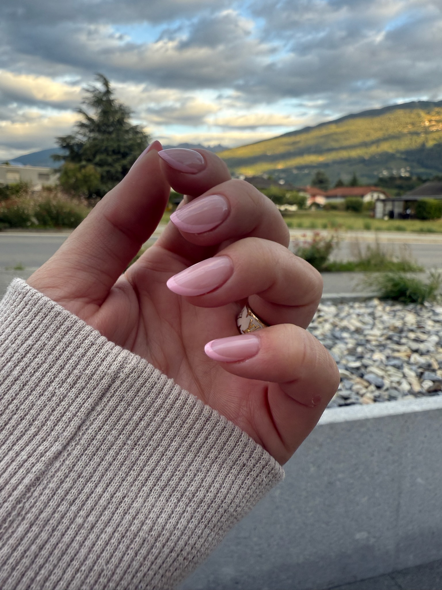 Une main aux ongles longs, brillants et manucurés en rose pâle est tenue à l'extérieur. La personne porte un pull beige à côtes. Des montagnes, des arbres, des nuages et une route sont visibles en arrière-plan.