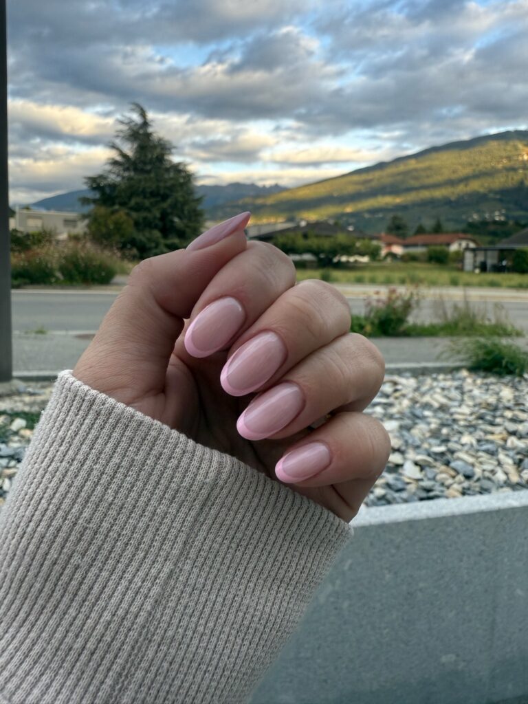 Une main aux ongles courts et ovales, de couleur rose pâle, est tenue à l'extérieur, une manche couvrant partiellement le poignet. À l'arrière-plan, on distingue des montagnes, un ciel nuageux, de la verdure et des rochers.