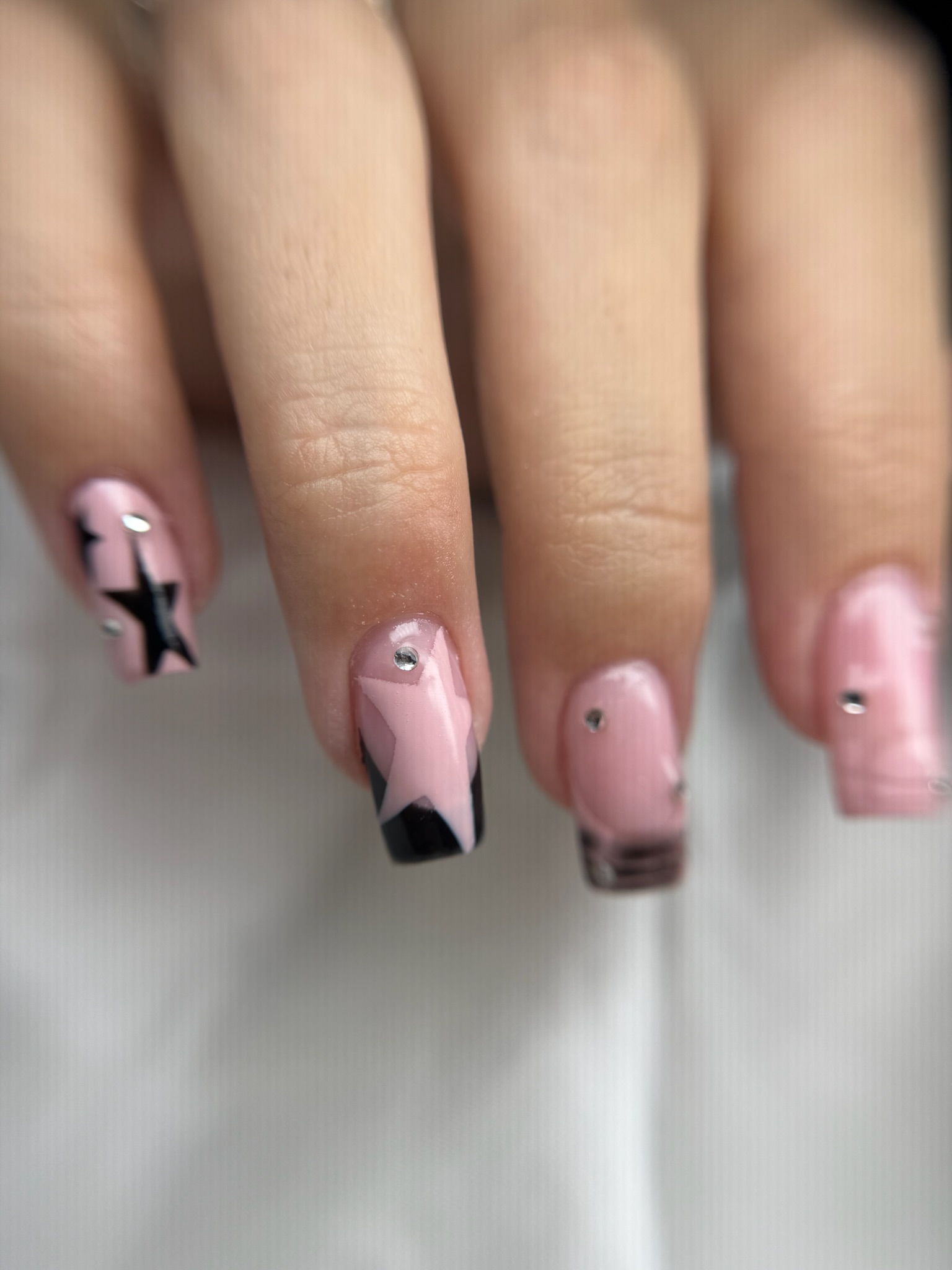 Gros plan sur une main aux ongles carrés peints en rose pâle, avec des motifs étoilés noirs et de petits strass argentés sur certains ongles. L'arrière-plan est flou, ce qui met en valeur le nail art.