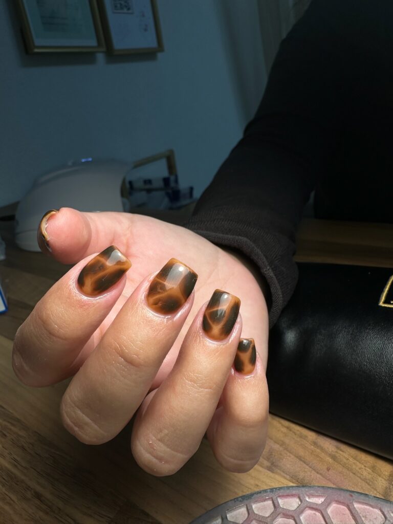 Une main aux ongles longs et carrés, ornés d'un motif brillant d'écailles de tortue brunes et ambrées, posée sur une table en bois sous une lumière vive.