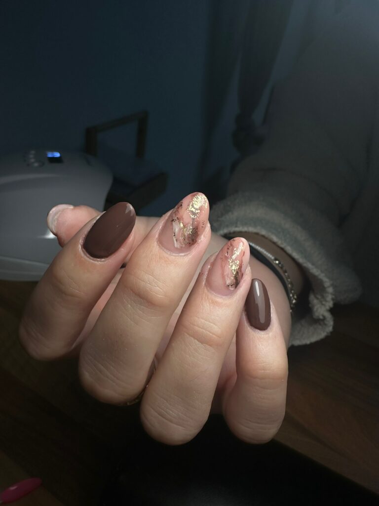Une main aux ongles manucurés présentant un mélange de vernis brun solide et de vernis transparent avec des accents de feuille d'or, montrée sous un éclairage doux sur une surface en bois.