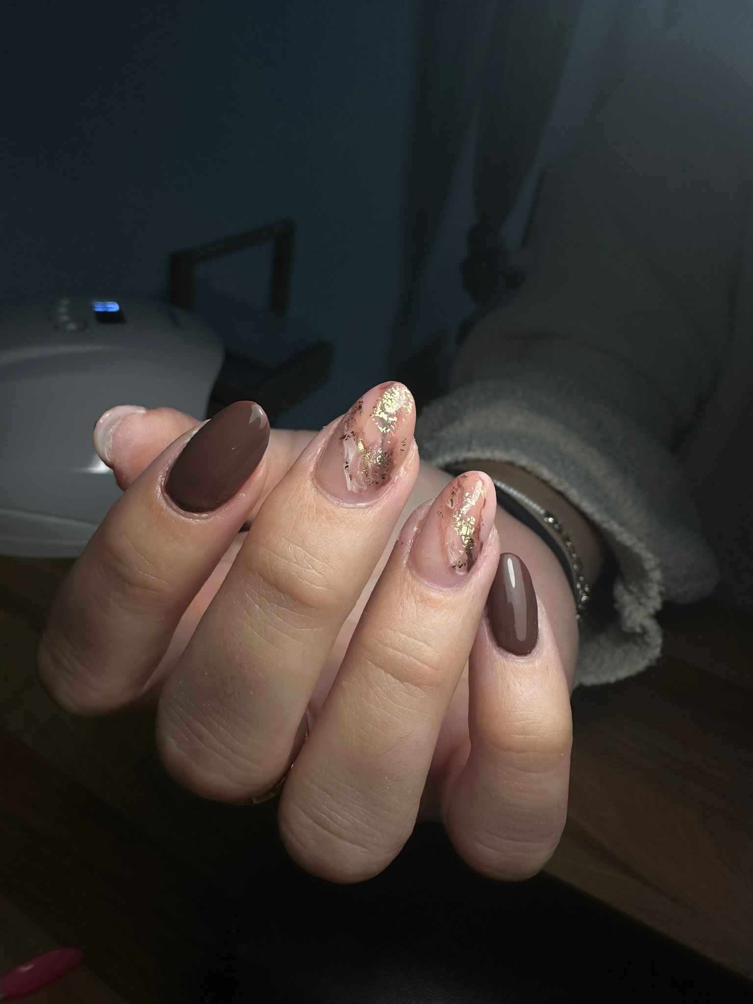 Une main aux ongles manucurés présentant un mélange de vernis brun solide et de vernis transparent avec des accents de feuille d'or, montrée sous un éclairage doux sur une surface en bois.