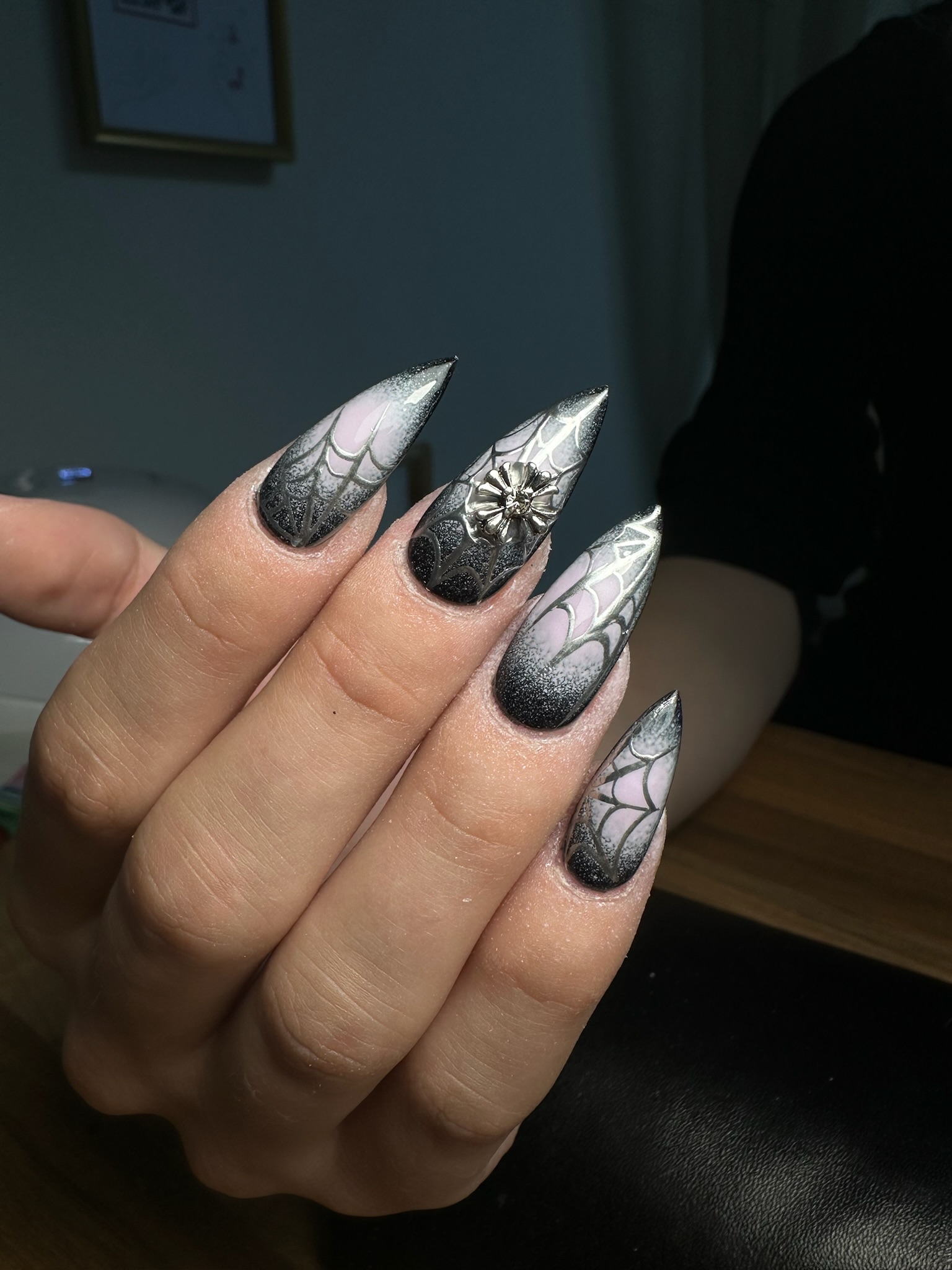 Une main aux ongles longs et pointus présentant des motifs de toile d'araignée noirs et blancs et une décoration d'araignée en 3D sur l'un des ongles, présentée sous une lumière vive.