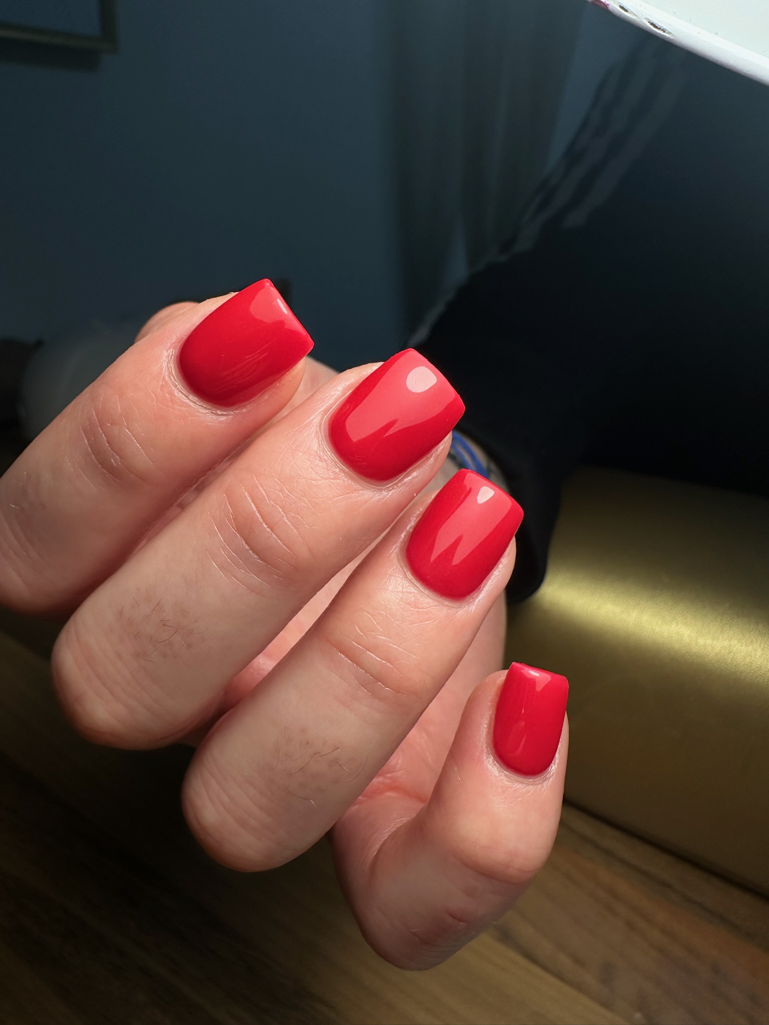 Gros plan d'une main aux ongles fraîchement manucurés, peints avec un vernis rouge brillant, dont les pointes sont de forme carrée. La main repose sur une surface en bois sous une lumière vive.